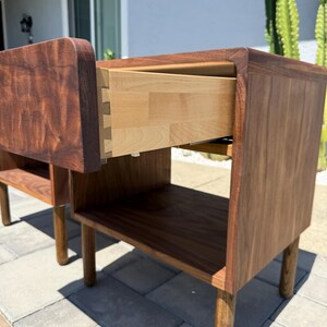 May include: A dark brown wooden nightstand featuring a single drawer with visible dovetail joinery and a light-colored interior. The piece has a smooth, curved top edge, a lower open storage shelf, and four cylindrical legs extending to the floor.