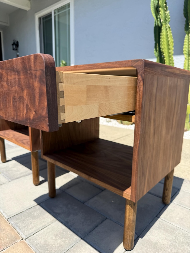 May include: A dark brown wooden nightstand featuring a single drawer with visible dovetail joinery and a light-colored interior. The piece has a smooth, curved top edge, a lower open storage shelf, and four cylindrical legs extending to the floor.