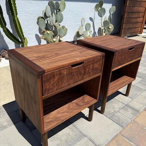 May include: A pair of matching dark brown wooden nightstands, each featuring a single top drawer with a recessed handle and an open storage shelf below. The nightstands are supported by four cylindrical legs and display a prominent wood grain.