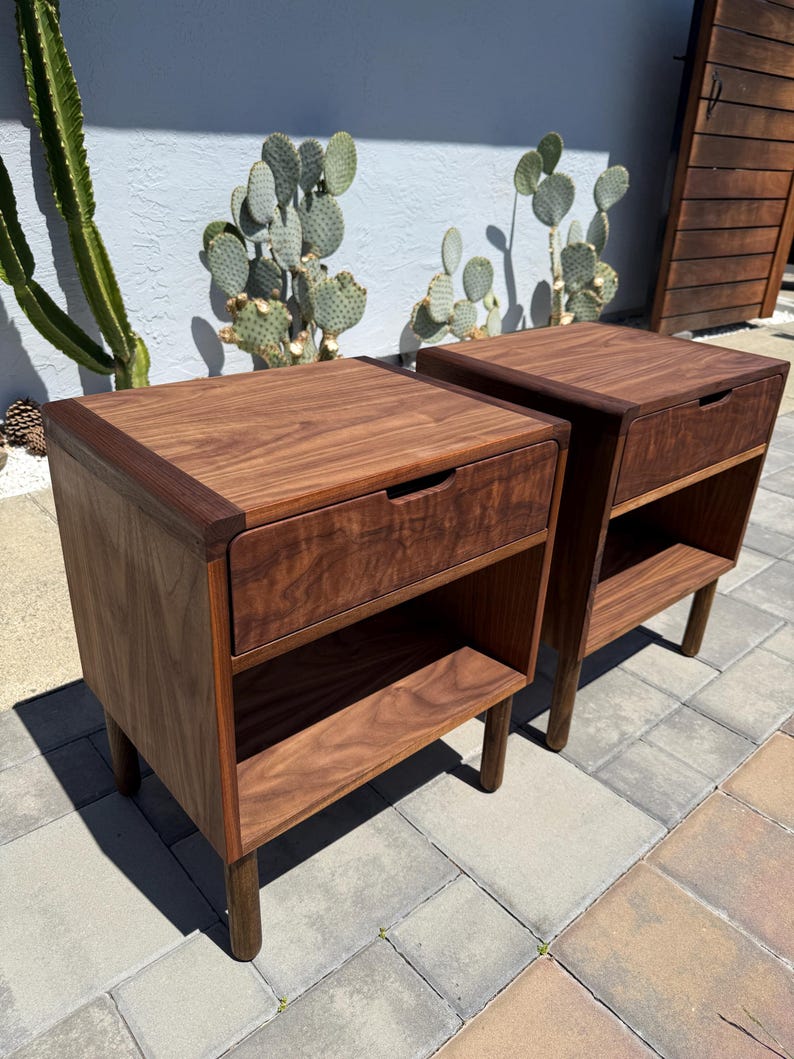 May include: A pair of matching dark brown wooden nightstands, each featuring a single top drawer with a recessed handle and an open storage shelf below. The nightstands are supported by four cylindrical legs and display a prominent wood grain.