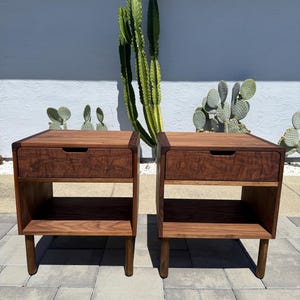 May include: A pair of matching wooden nightstands, each featuring a single top drawer with a recessed handle, an open storage shelf below, and four cylindrical legs. The furniture is constructed from dark brown wood with visible grain patterns.