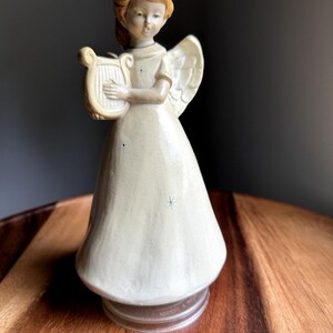 Può includere: Statuina di angelo in ceramica con abito e ali color crema, che tiene un'arpa. L'angelo ha i capelli castano chiaro ed è decorato con piccoli accenti di stelle blu. La statuina è posta su una base marrone e su una superficie di legno.