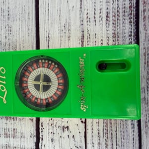 May include: A bright green rectangular plastic game device featuring a circular roulette-style wheel with numbers and a gold-colored center spinner. The text Lotto is printed in gold on the left, and Spin-A-Winner is printed in gold on the right.