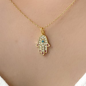 May include: Gold necklace with a hamsa hand pendant featuring a turquoise evil eye and pink and white cubic zirconia stones.