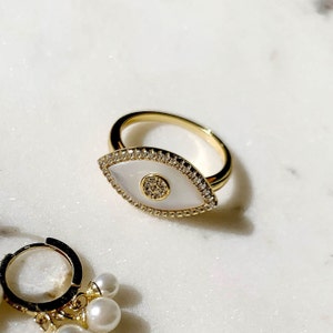May include: A gold ring with a white evil eye design. The eye is surrounded by small clear stones.