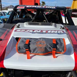 May include: A white, red, and black off-road racing vehicle with the word "CANTINA" across the top. A gray bag with orange straps and the text "SURVIVAL OUTDOOR SYSTEMS" is secured on the hood.