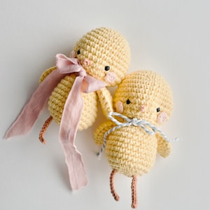 May include: Two crocheted yellow chick toys with embroidered eyes and beaks. One chick has a pink ribbon tied around its neck, and the other has a blue ribbon.
