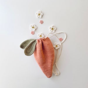 Puede incluir: Una bolsa de cordón de crochet con forma de zanahoria con hojas verdes, decorada con flores de crochet blancas y rosas.