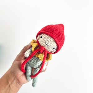 Puede incluir: Muñeca de ganchillo hecha a mano con un gorro puntiagudo rojo, un cuello con volantes amarillos y lazos rojos. La muñeca tiene un cuerpo gris claro y una cara sonriente. La muñeca se sostiene en una mano sobre un fondo blanco.