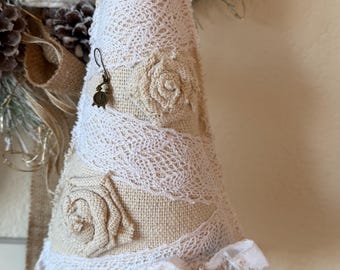 May include: A cone-shaped decoration made of beige burlap, wrapped in white lace ribbons and adorned with fabric rosettes. A small bronze-colored clock charm hangs from the side, and a white ribbon bow with a heart pattern is tied at the base.