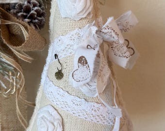 May include: A cone-shaped fabric ornament in cream burlap, decorated with white lace trim, fabric roses, and a white ribbon featuring a printed heart and text. A small metal safety pin charm with the words Made with Love hangs on the front.