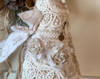 May include: A fabric tree ornament in beige and white, decorated with intricate lace, two frayed fabric rosettes, and a small heart-shaped metal charm inscribed with the words "made with love" attached by a safety pin, featuring a white stand.