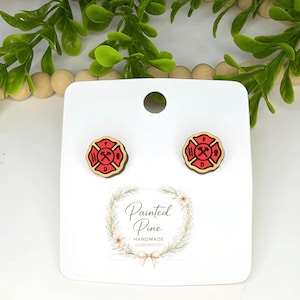 May include: Wooden stud earrings featuring a red and black firefighter Maltese cross design. The earrings are on a white card with the text "Painted Pine Handmade" and a floral wreath.