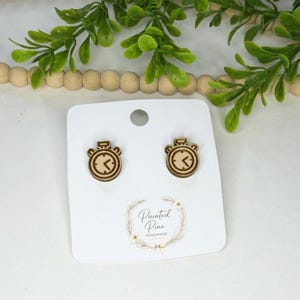 May include: Wooden clock-shaped stud earrings. The earrings are light brown with a clock face design. The earrings are displayed on a white card with the text "Painted Pine Handmade". Greenery and wooden beads are in the background.