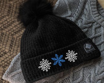 May include: Black knitted beanie with a faux fur pom pom and embroidered snowflakes. The beanie has a Columbia logo patch on the side.
