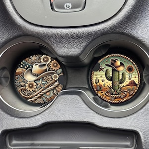 May include: Two round car coasters with a western theme. One coaster features a cowboy hat with stars and flowers. The other coaster features a cactus with a cowboy hat and a lasso.
