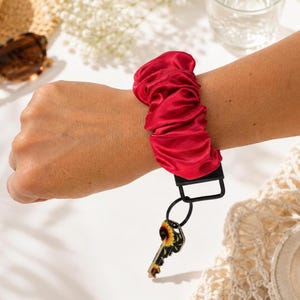 May include: A red satin scrunchie wristlet featuring a black metal clasp and a black key ring attached to a single key with a sunflower pattern. The fabric has a gathered, pleated texture and a smooth, reflective finish on the surface.