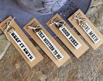 May include: Four keychains with white fabric straps and black text. The phrases read "KEEP IT CUTE", "TALK WESTERN TO ME", "GOOD VIBES", and "GIRL MATH". Each keychain is attached to a brown card with a metal clasp and white string. A gray hat with floral design is in the background.