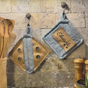May include: Two handmade pot holders hanging on hooks. One is tan with gray accents and grommets. The other is blue with a tan patch that reads "Edwards". Both have a diamond shape and are made of fabric.