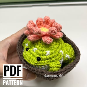 May include: A crocheted cactus with a pink flower on top, sitting in a brown pot. The cactus is green and has white accents. The pot is brown and has a crocheted texture. The image includes the text "PDF PATTERN" and "@yarnieayarniecrochet".