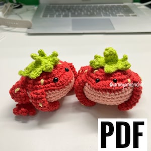 Puede incluir: Dos peluches de rana de fresa de crochet con hojas verdes en la parte superior. Los peluches son rojos y rosados con ojos negros. La imagen incluye el texto "PDF PATTERN".