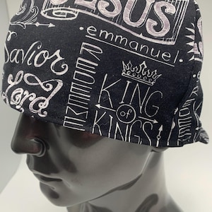 May include: Black cap with white script and illustrations. The cap features the word "JESUS" in large letters, along with other religious phrases and a crown graphic. The cap is displayed on a mannequin head.