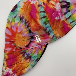 May include: A colorful tie-dye welding cap with a black brim. The cap features a vibrant mix of pink, orange, yellow, green, and purple hues. A small white tag with black text is attached to the cap.