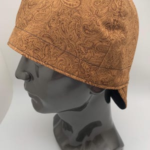 May include: A brown paisley patterned welding cap with a black leather flap. The cap is worn on a black mannequin head.