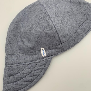 May include: A gray paisley print welding cap. The cap features a curved brim and is made of a soft fabric. The paisley pattern is a lighter shade of gray against a darker gray background. A small tag is attached to the brim.