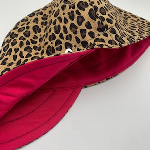 May include: A brown and black leopard print fabric hat with a bright pink lining. The hat has a button on the top.