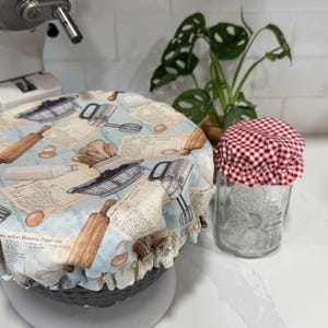 May include: A light blue and beige fabric bowl cover with a vintage baking-themed print, covering a white stand mixer. A glass jar with a red and white checkered lid sits nearby. A green plant is in the background.