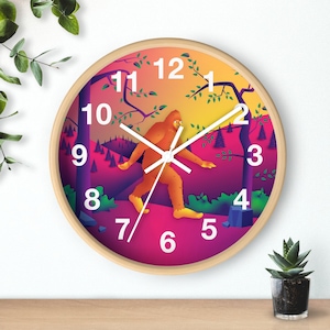 May include: A round wall clock with a colorful design featuring a cartoon Bigfoot walking through a forest. The clock face has a pink, orange, and yellow gradient background with white numbers and hands.