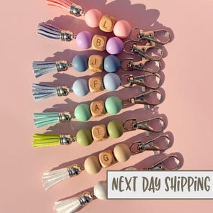 May include: A collection of beaded keychains in various pastel colors, each featuring a tassel and a metal clasp. The keychains have wooden lettered beads spelling out individual names. The text "NEXT DAY SHIPPING" is visible.