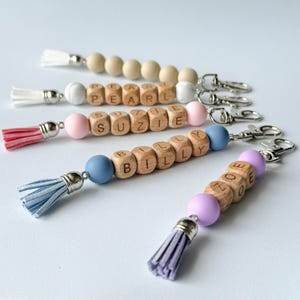 May include: Five personalized keychains featuring wooden beads with engraved names including Pearl, Suzie, Billy, and Zoe. Each keychain includes colorful silicone beads, a matching faux suede tassel, and a silver-toned metal lobster clasp.