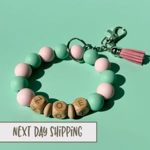 May include: A light pink and mint green silicone beaded keychain with a wooden charm that says "ZOE" and a pink tassel.