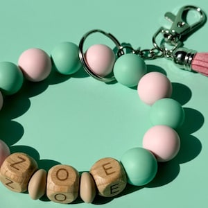 May include: A circular keychain featuring alternating matte pastel pink and mint green silicone beads. Three light brown wooden cube beads are engraved with the letters Z, O, and E. A silver metal keyring and lobster clasp are attached with a pink tassel.