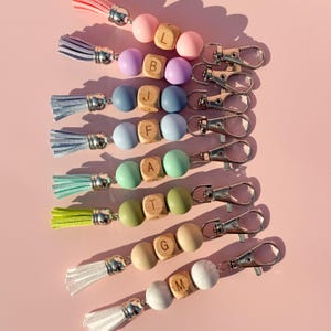 May include: A collection of eight keychains featuring colorful tassels, two round silicone beads, and a central wooden cube bead engraved with a letter. Each keychain is attached to a silver-toned metal lobster clasp, showcasing a variety of pastel and vibrant hues including pink, purple, blue, green, yellow, beige, and white.