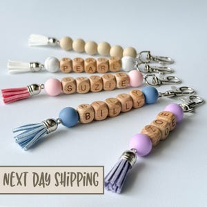 May include: Five personalized wooden bead keychains with different names, each with a colorful silicone bead and a tassel. The names are Pearl, Suzie, Billy, and Zoe.