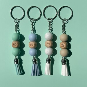 May include: Four keychains featuring a metal ring and chain, each with two round beads, one wooden bead engraved with a letter, and a faux leather tassel. The beads are mint green, light blue, white marble, and beige, with matching tassels.