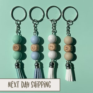 May include: Four keychains with different colored beads and wooden letter charms. The beads are light blue, light green, white, and beige. The charms are engraved with the letters S, C, J, and M. Each keychain has a silver tassel.