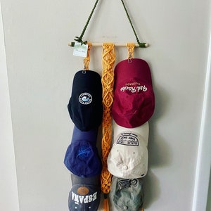 May include: A macrame wall hanging with five baseball caps hanging from it. The caps are in various colors and have different logos and designs. The macrame is made of yellow cord and has a wooden dowel at the top.