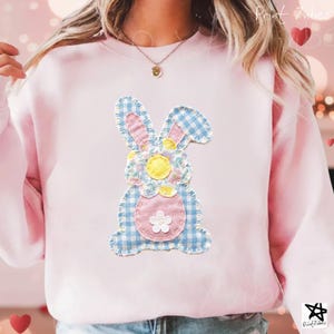 May include: A light pink sweatshirt featuring a patchwork bunny design. The bunny has blue gingham ears and body, a pink belly, and a yellow flower. The design is stitched onto the sweatshirt, creating a textured look.