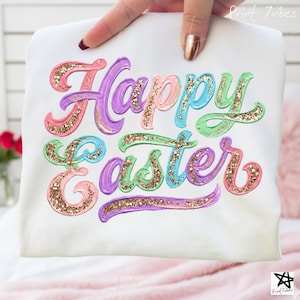May include: White t-shirt with the words "Happy Easter" in a colourful, glittery design. The text features pink, purple, blue, and green hues with gold glitter accents. The shirt is held up by a hand.