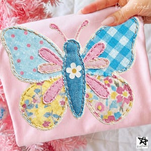 Könnte beinhalten: Ein rosa Stoffartikel mit einem großen, farbenfrohen Schmetterlingsaufnäher. Der Schmetterling hat Flügel aus blauem Tupfen-, Vichy- und Blumenmusterstoffen. Der Schmetterlingskörper ist aus Jeans mit einer weißen Blume. Der Text "Print 7vibes" ist sichtbar.