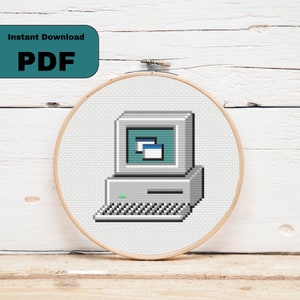 Puede incluir: Un patrón de punto de cruz de una computadora vintage con un diseño pixelado. La computadora es gris con una pantalla azul y una ventana blanca abierta en la pantalla. La computadora tiene un teclado y una unidad de disquete. El patrón está en un aro de bordado redondo.