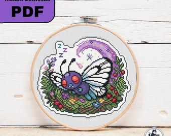 Op de afbeelding: Een kruissteekpatroon in een houten borduurring met een paars insect met grote rode ogen en witte vleugels dat slaapt op een bed van groen gebladerte, kleine kleurrijke bloemen en rode bessen onder een paarse maansikkel met ZZZ-tekst.
