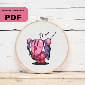 May include: A cross-stitch pattern featuring a pink Kirby character singing with a conductor's baton. The pattern is in a white embroidery hoop.