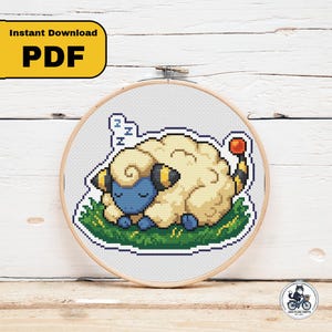 Könnte beinhalten: Ein Kreuzstichmuster in einem hölzernen Stickrahmen mit einem Pixel-Art-Design eines schlafenden, schafähnlichen Wesens mit cremefarbener Wolle, blauem Gesicht und einem gestreiften Schwanz mit roter Kugelspitze, das auf grünem Gras ruht.