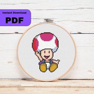 Könnte beinhalten: Kreuzstichmuster eines roten und weißen Pilzcharakters mit einem lila Hemd und gelben Schuhen. Das Muster wird in einem hölzernen Stickrahmen angezeigt.