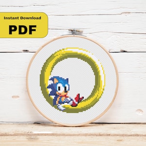 May include: Cross-stitch embroidery of Sonic the Hedgehog inside a yellow ring. The design is set in a wooden embroidery hoop. The text "Instant Download PDF" is in a yellow rectangle in the upper left corner.